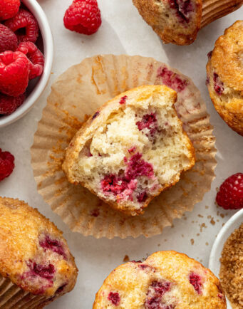 A partially eaten raspberry muffin sits on an open wrapper, surrounded by whole muffins and fresh raspberries on a light surface.