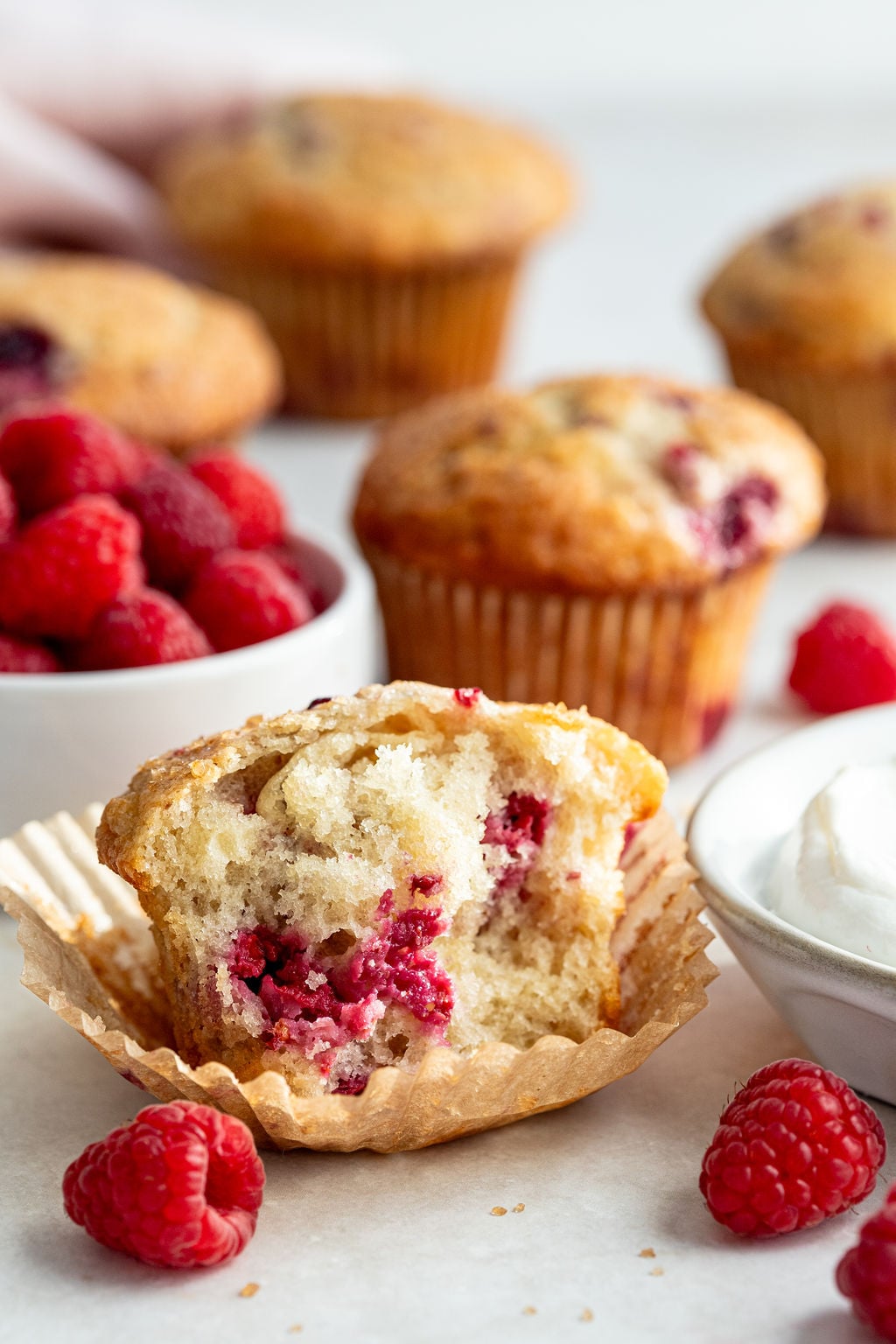A raspberry muffin with a bite taken out sits in a wrapper, surrounded by fresh raspberries, whole muffins, and a bowl of cream on a light surface.