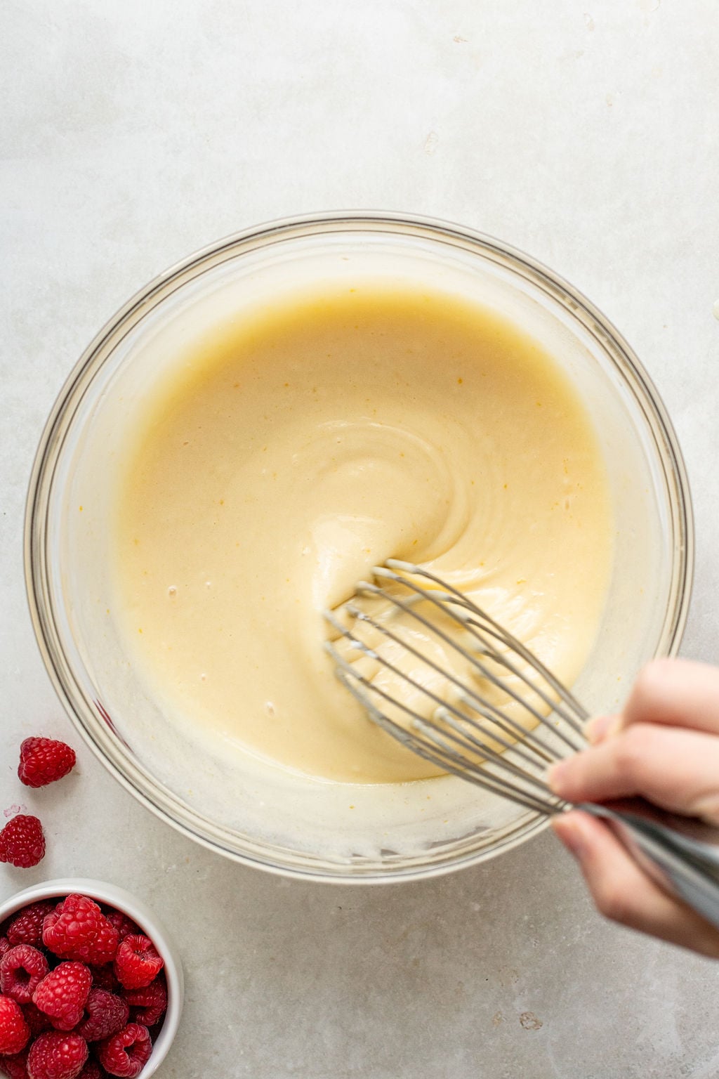 A hand whisking a smooth, pale batter in a glass bowl, with a small bowl of fresh raspberries nearby on a light countertop.
