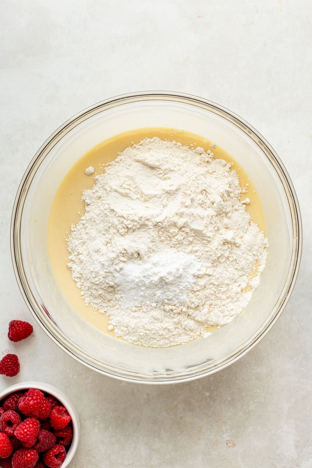 A glass bowl with flour and baking powder on top of a yellow batter, next to a small bowl of fresh raspberries on a white surface.