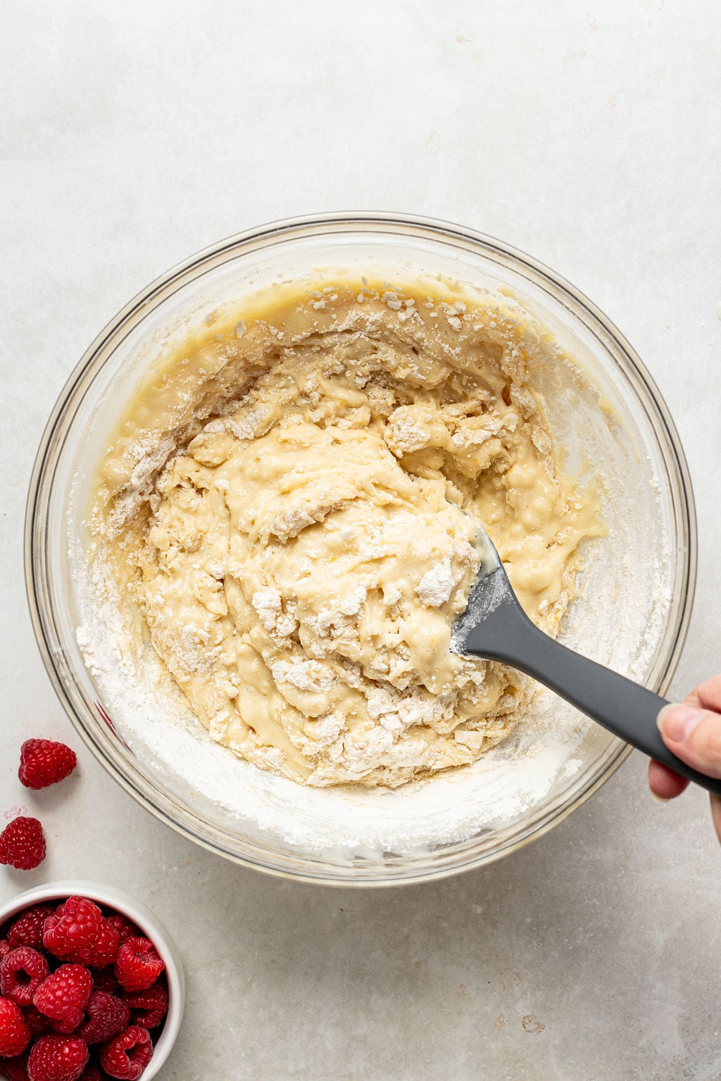 A hand mixes thick batter in a glass bowl with a black spatula; fresh raspberries are in a small white bowl nearby on a white surface.