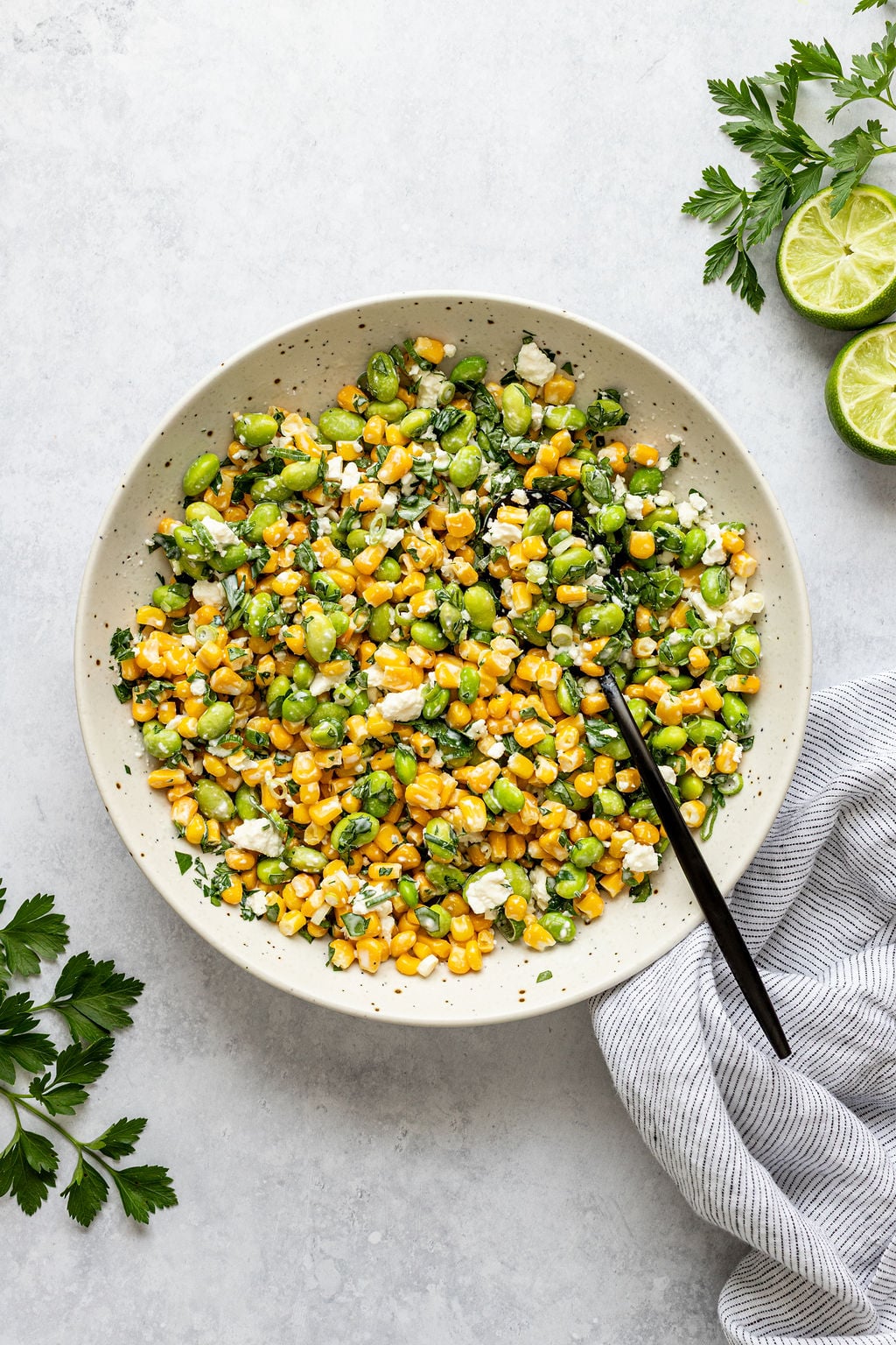 A bowl of corn and edamame salad with herbs and feta cheese sits on a light surface, surrounded by parsley, lime slices, and a striped napkin.