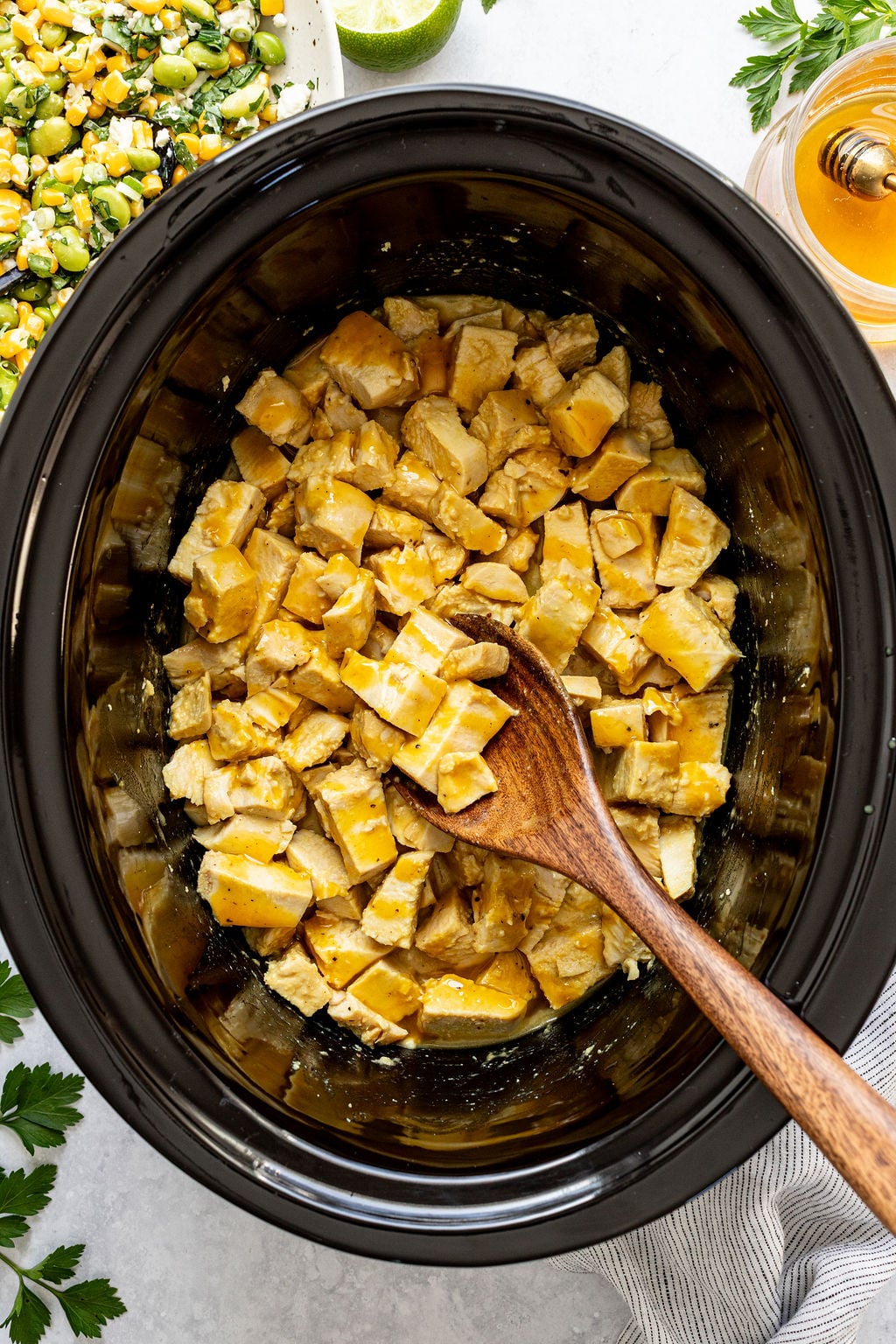 Cubed cooked chicken in a slow cooker with a wooden spoon, drizzled with sauce. A plate of mixed corn and herbs is partially visible nearby.