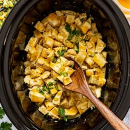 Diced cooked chicken with sauce, garnished with chopped green onions and parsley, in a black slow cooker with a wooden spoon.