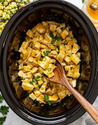 Diced cooked chicken with sauce, garnished with chopped green onions and parsley, in a black slow cooker with a wooden spoon.