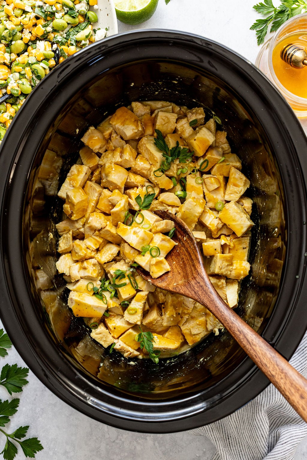 Diced cooked chicken with sauce, garnished with chopped green onions and parsley, in a black slow cooker with a wooden spoon.