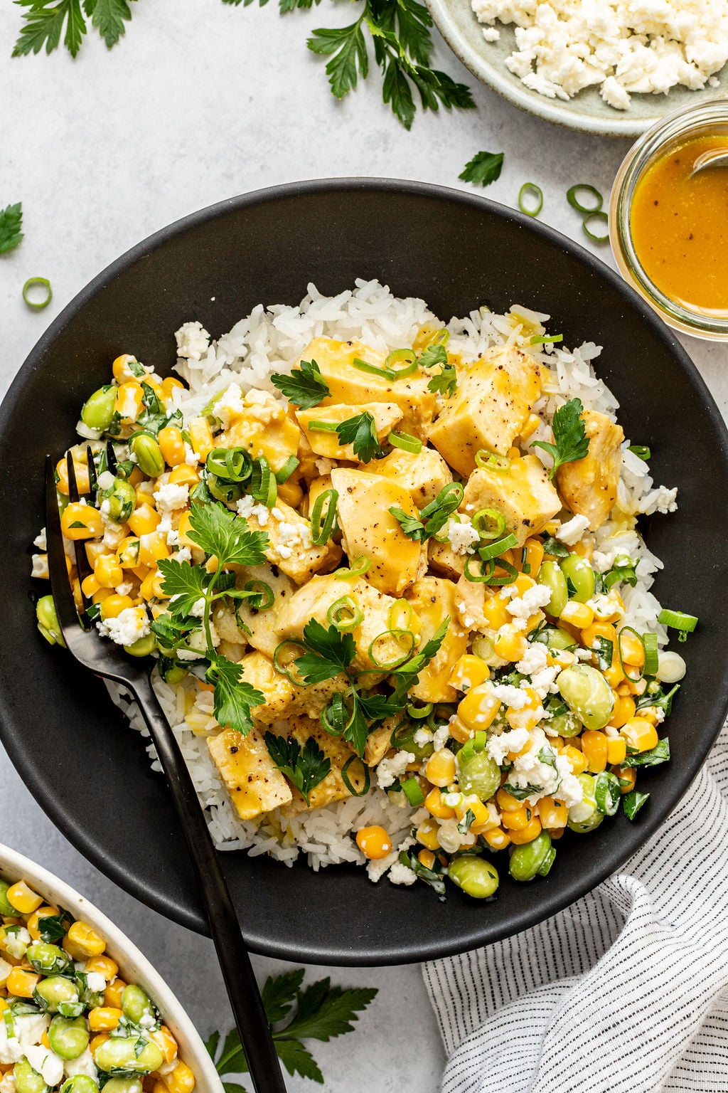 A black bowl filled with white rice topped with seasoned chicken, corn, edamame, green onions, cilantro, and crumbled cheese. A fork rests in the bowl.