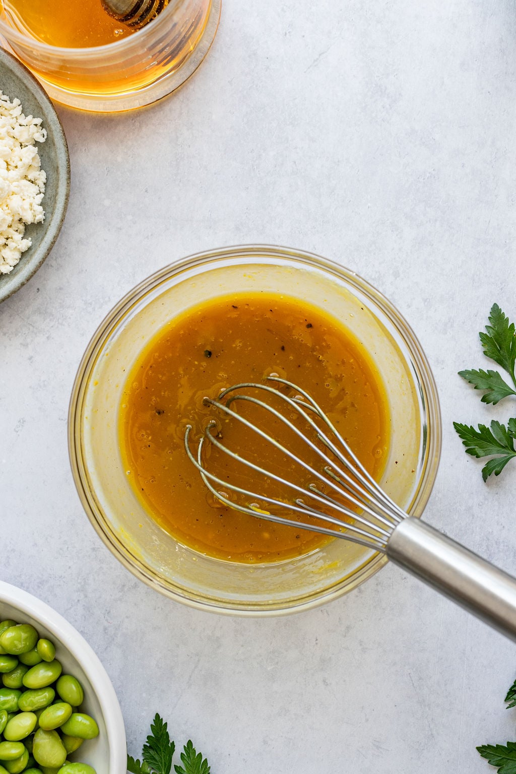 A glass bowl of yellow dressing being whisked, surrounded by honey, cheese, parsley, and a bowl of green edamame on a light surface.