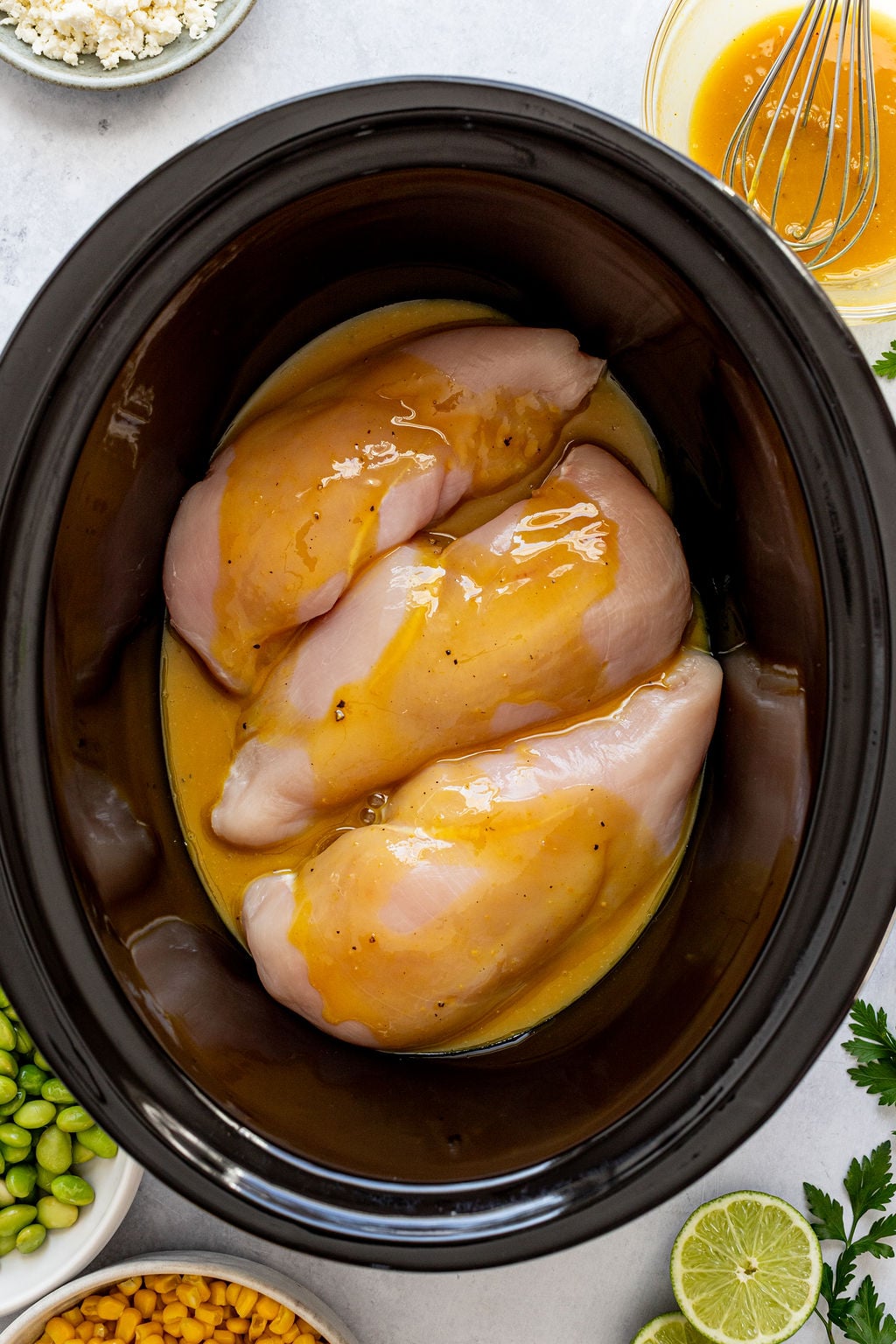 Three raw chicken breasts coated in sauce inside a slow cooker, surrounded by bowls of ingredients including edamame, corn, lime slices, and a whisked sauce.