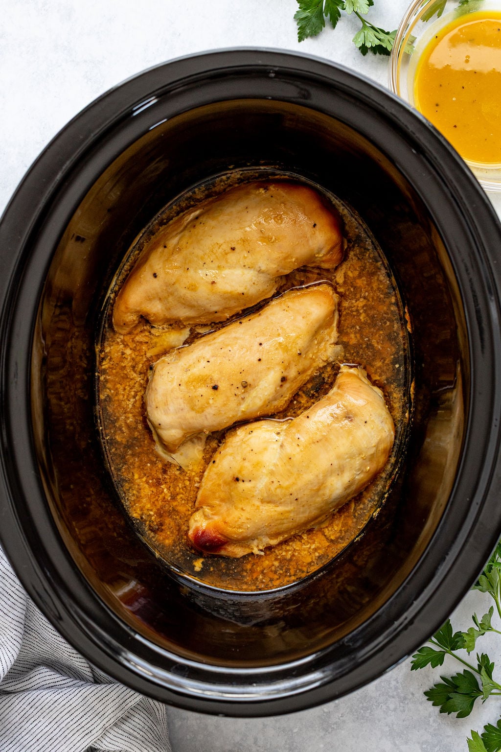 Three cooked chicken breasts in sauce sit inside a black slow cooker, with fresh herbs and a bowl of yellow sauce nearby on a light countertop.