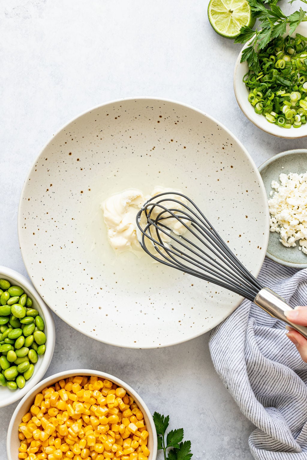 A whisk is mixing mayonnaise in a large speckled bowl, surrounded by small bowls of corn, edamame, green onions, feta cheese, and herbs.