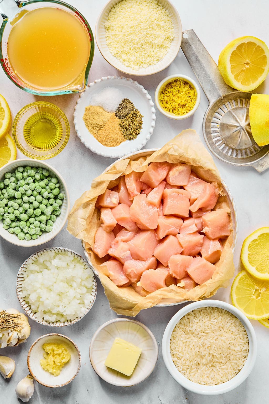 Top-down view of raw diced chicken, rice, frozen peas, chopped onions, broth, butter, lemon, spices, garlic, and grated cheese arranged in bowls and plates on a white surface.