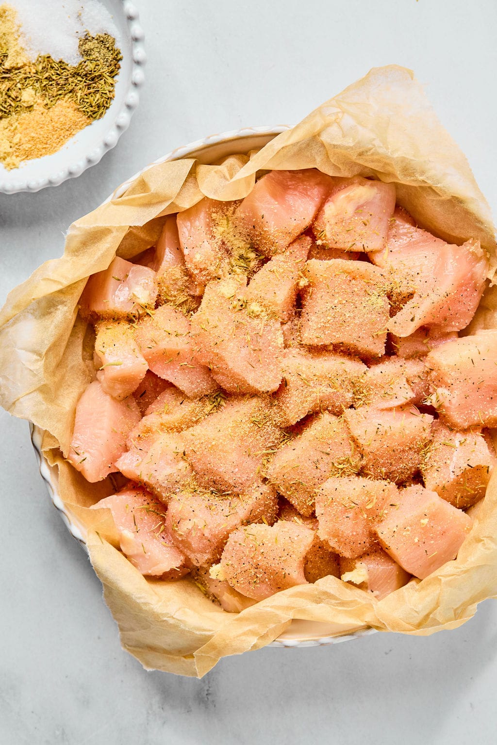 Chunks of raw chicken breast in a parchment-lined dish, sprinkled with dry seasoning; a bowl of additional seasoning is nearby on the countertop.