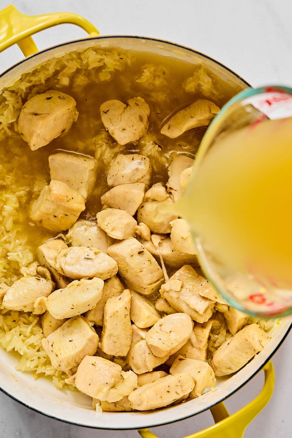 Cooked chicken pieces and rice in a yellow pot, with broth being poured from a measuring cup into the mixture.