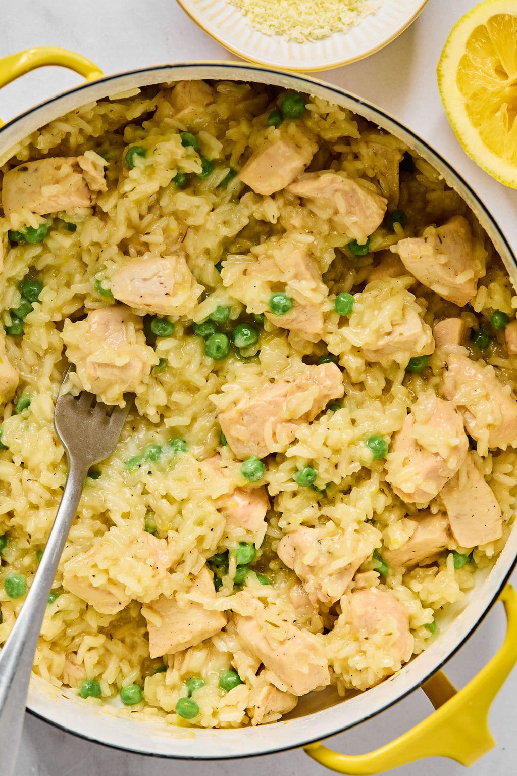 A pot of creamy rice with chunks of chicken and green peas, with a fork in the dish. A lemon wedge and a bowl of grated cheese are nearby.