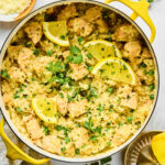 A pot of creamy lemon chicken and rice garnished with lemon slices and fresh parsley, with parmesan cheese and lemon slices on the side.