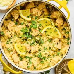 A pot of creamy lemon chicken and rice garnished with lemon slices and fresh parsley, with parmesan cheese and lemon slices on the side.