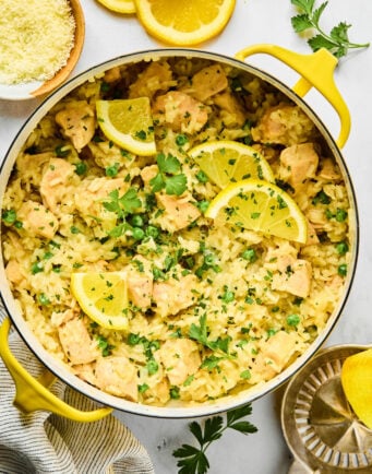 A pot of creamy lemon chicken and rice garnished with lemon slices and fresh parsley, with parmesan cheese and lemon slices on the side.
