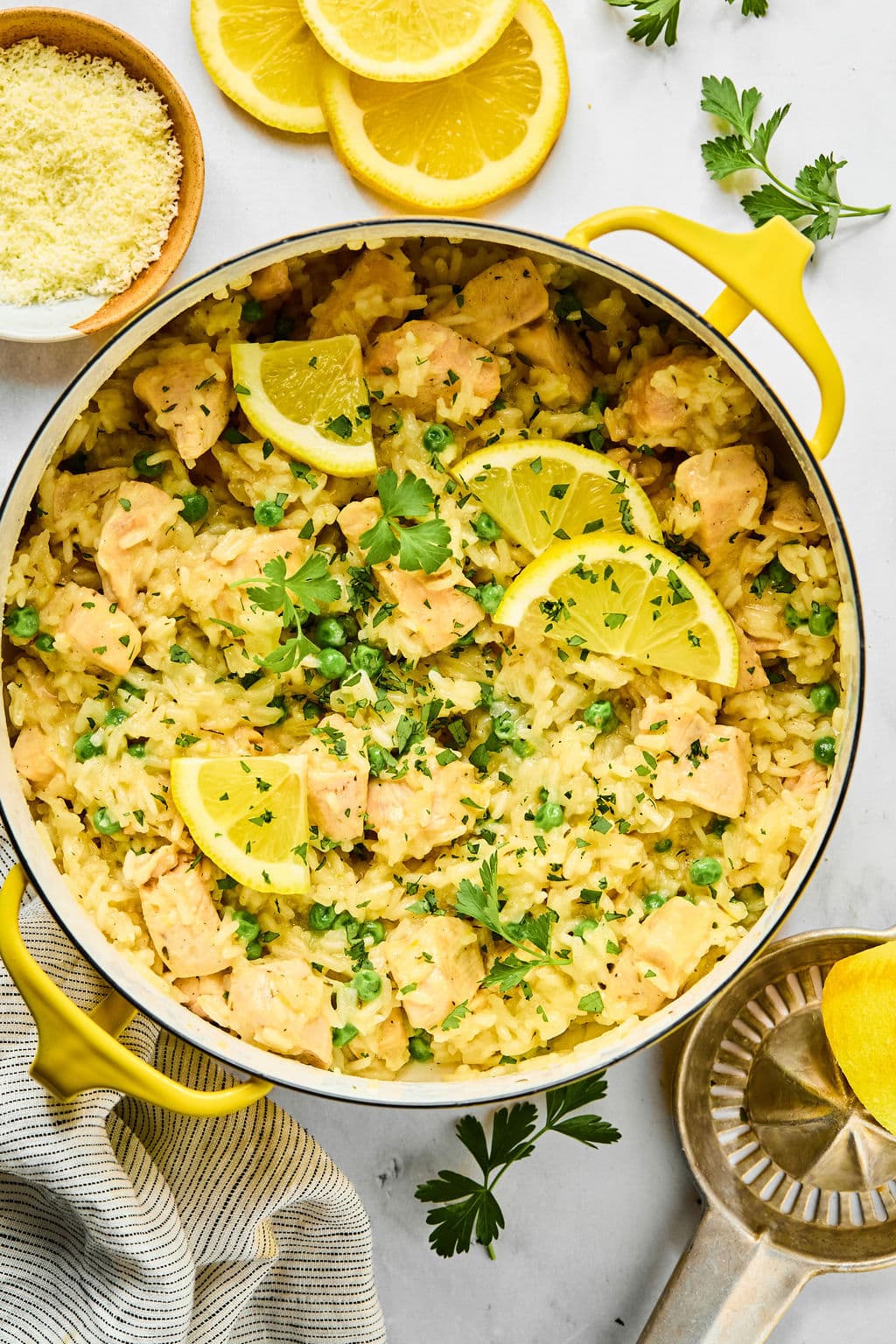 A pot of creamy lemon chicken and rice garnished with lemon slices and fresh parsley, with parmesan cheese and lemon slices on the side.