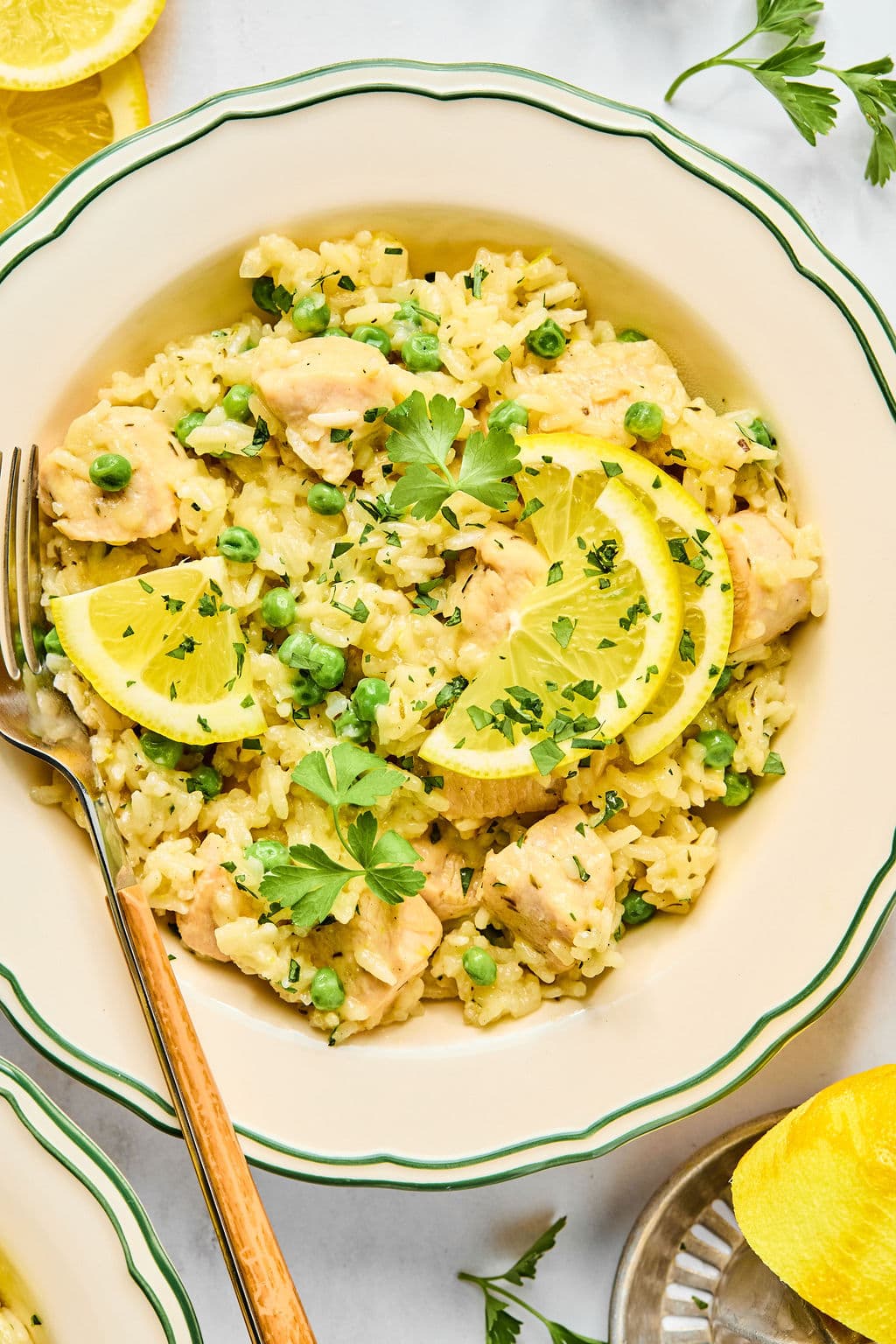 A bowl of chicken and pea risotto garnished with lemon slices and parsley, with a fork resting on the side.