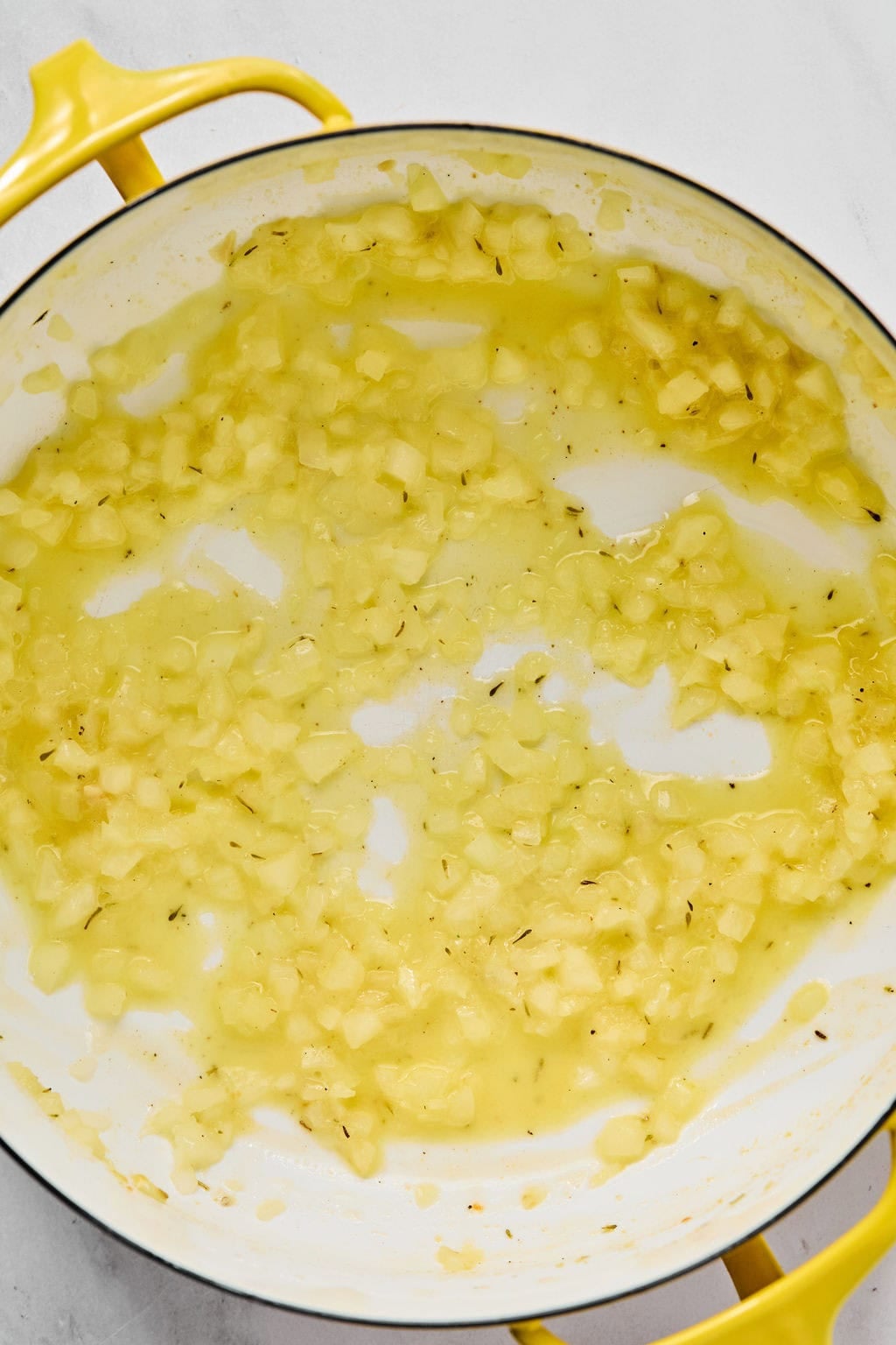 Chopped onions saut&eacute;ing in oil in a yellow-handled pan on a white surface.