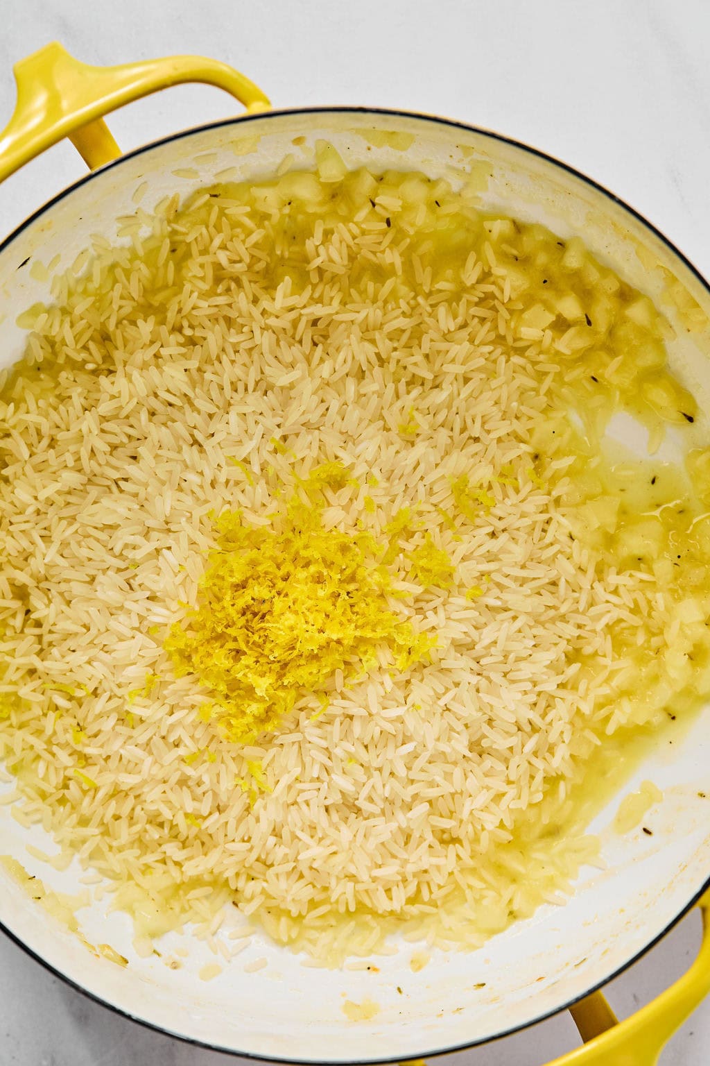 Uncooked rice and lemon zest are added to saut&eacute;ed onions in a yellow pot on a white surface.