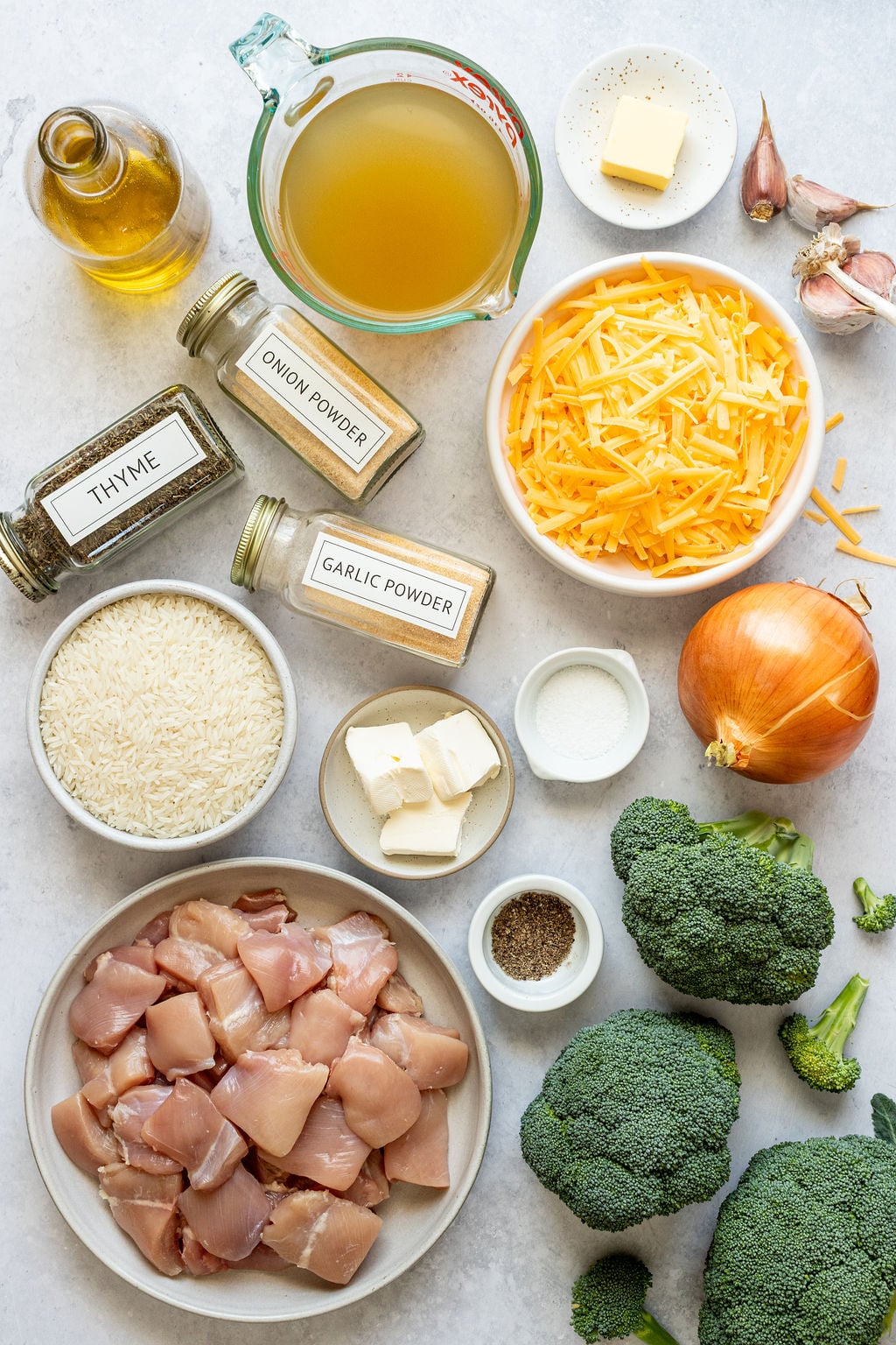 Ingredients for a chicken and broccoli casserole arranged on a countertop, including chicken, broccoli, rice, cheese, onion, spices, broth, butter, and oil.