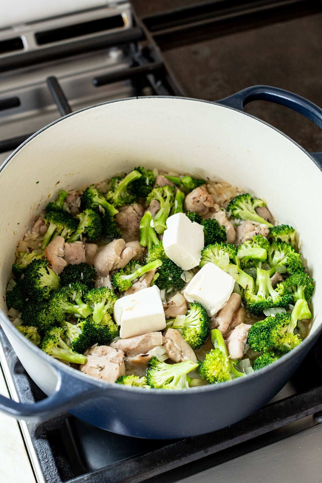 A pot on a stovetop containing chopped broccoli, pieces of cooked chicken, and several chunks of cream cheese.
