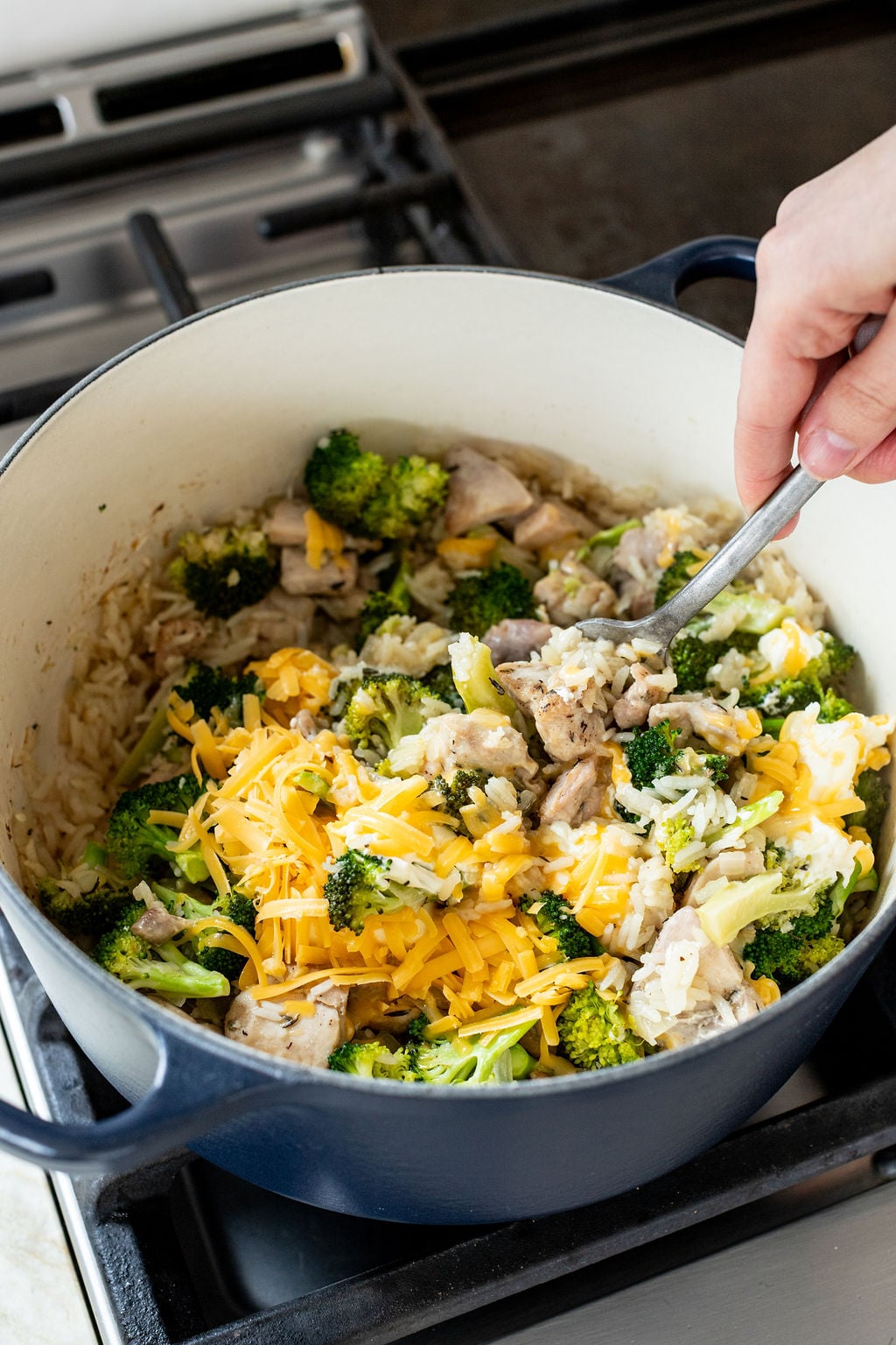A hand stirs a mixture of rice, broccoli, chicken, and shredded cheddar cheese in a large pot on a stove.