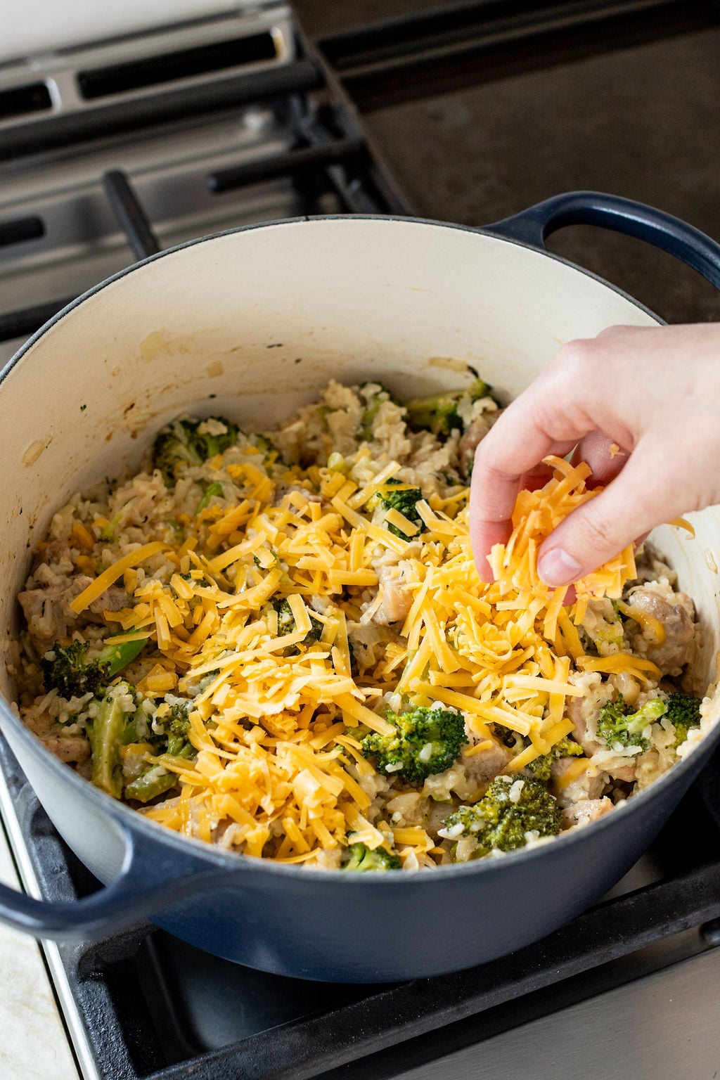 A hand sprinkles shredded cheddar cheese over a mixture of rice, broccoli, and chicken in a blue pot on a stovetop.