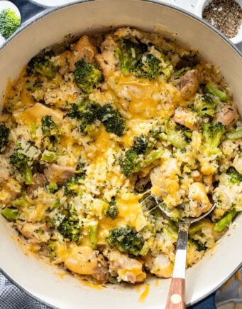 A pot filled with a broccoli, rice, and chicken casserole topped with melted cheddar cheese, with a serving spoon partially submerged.