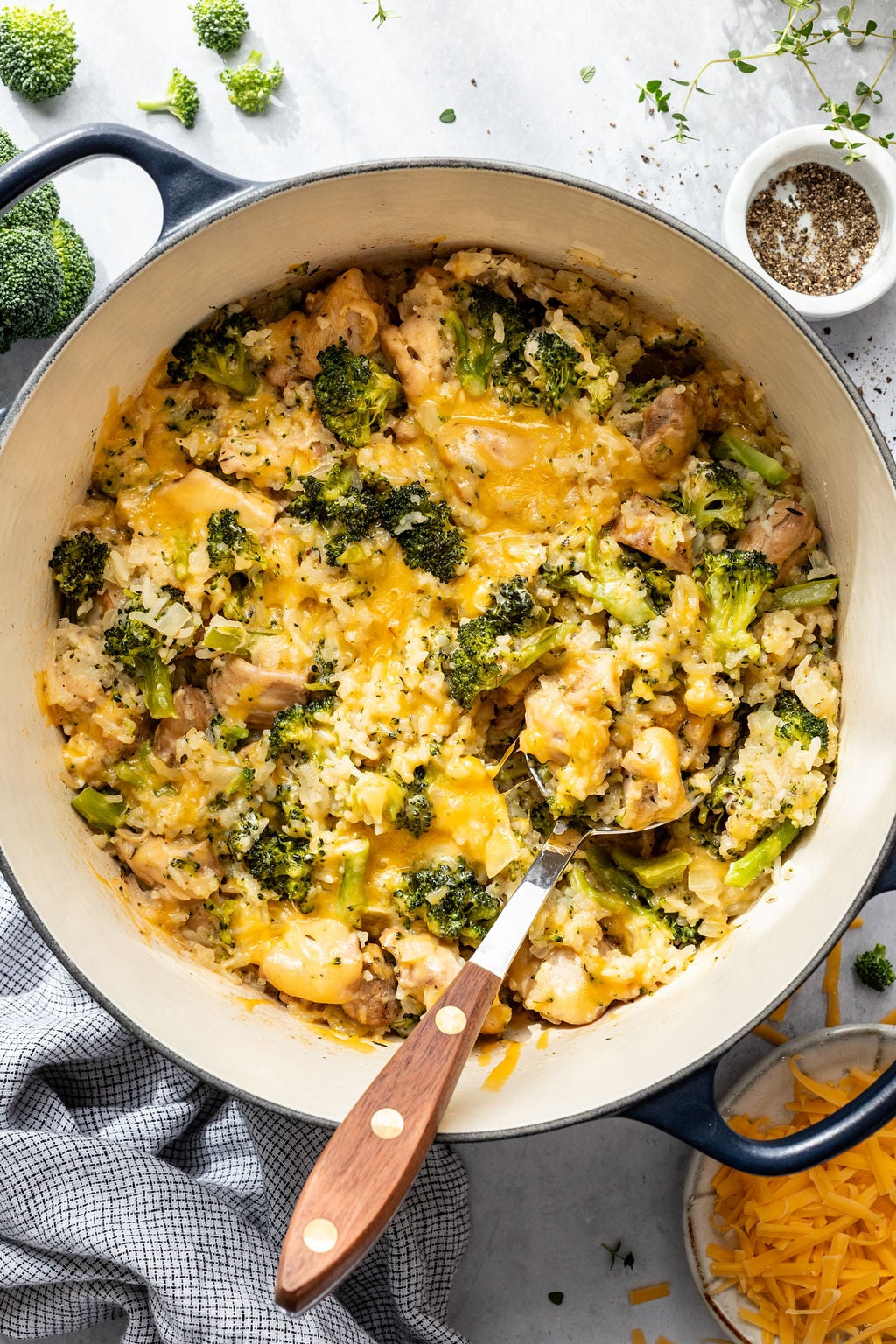 A large pot filled with cooked broccoli, rice, chicken, and melted cheese, with a serving spoon inside. Shredded cheese, pepper, and broccoli florets are nearby on the table.