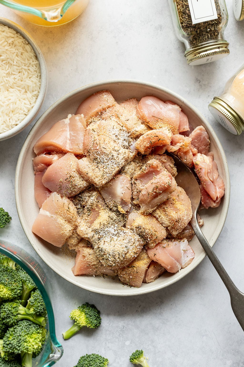 A bowl of raw, cubed chicken coated with dry seasoning sits on a countertop, surrounded by uncooked rice, broccoli, and spice jars.