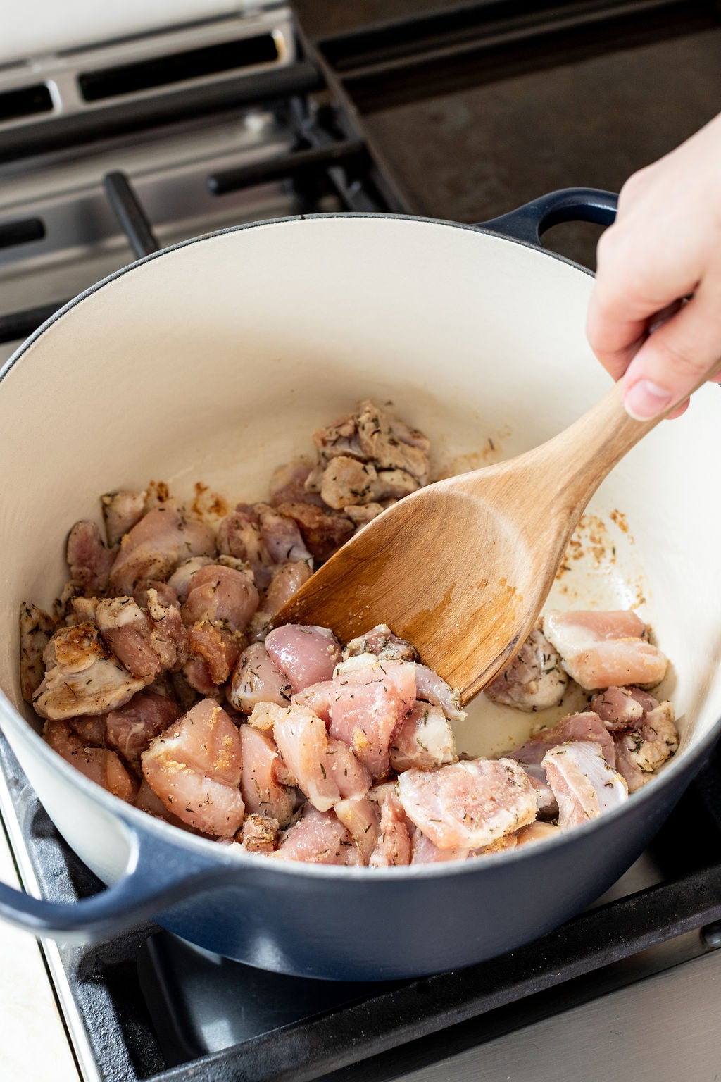 Chunks of raw chicken are stirred with a wooden spoon in a large white Dutch oven on a stovetop.