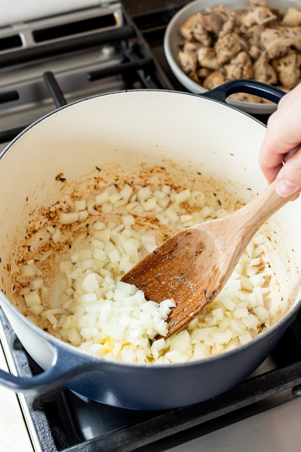 A hand stirs chopped onions in a large pot with a wooden spatula; cooked chicken pieces are on a plate in the background.