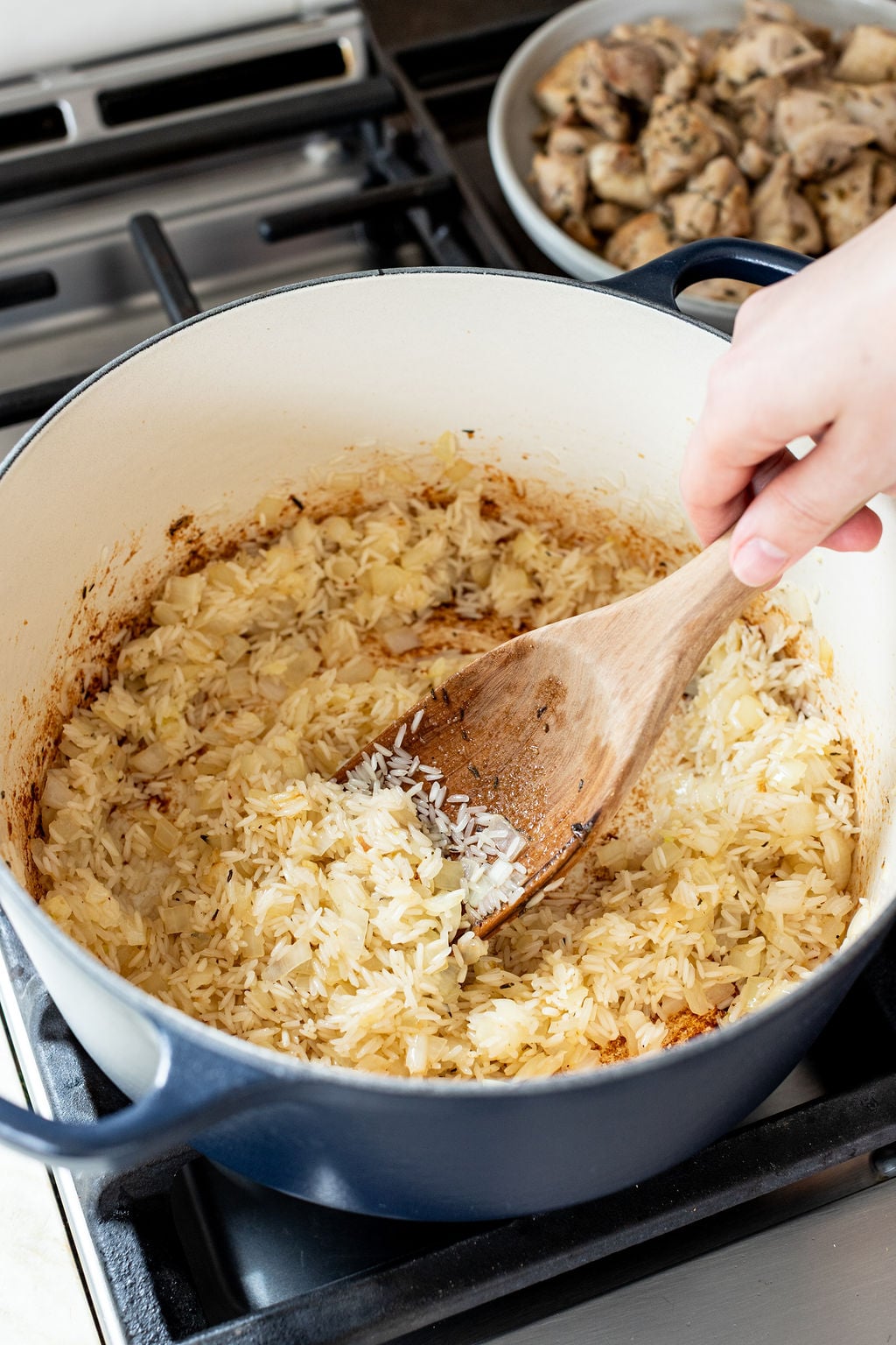 A hand stirs rice and saut&eacute;ed onions in a large pot with a wooden spoon; cooked chicken pieces are in a pan in the background on the stove.