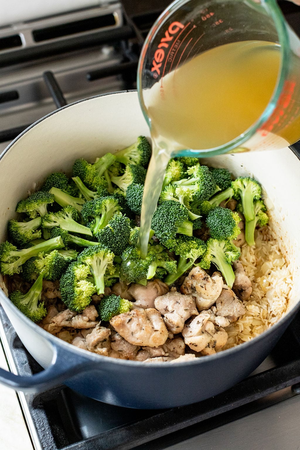 A person pours broth from a measuring cup into a pot containing broccoli, mushrooms, and rice on a stovetop.