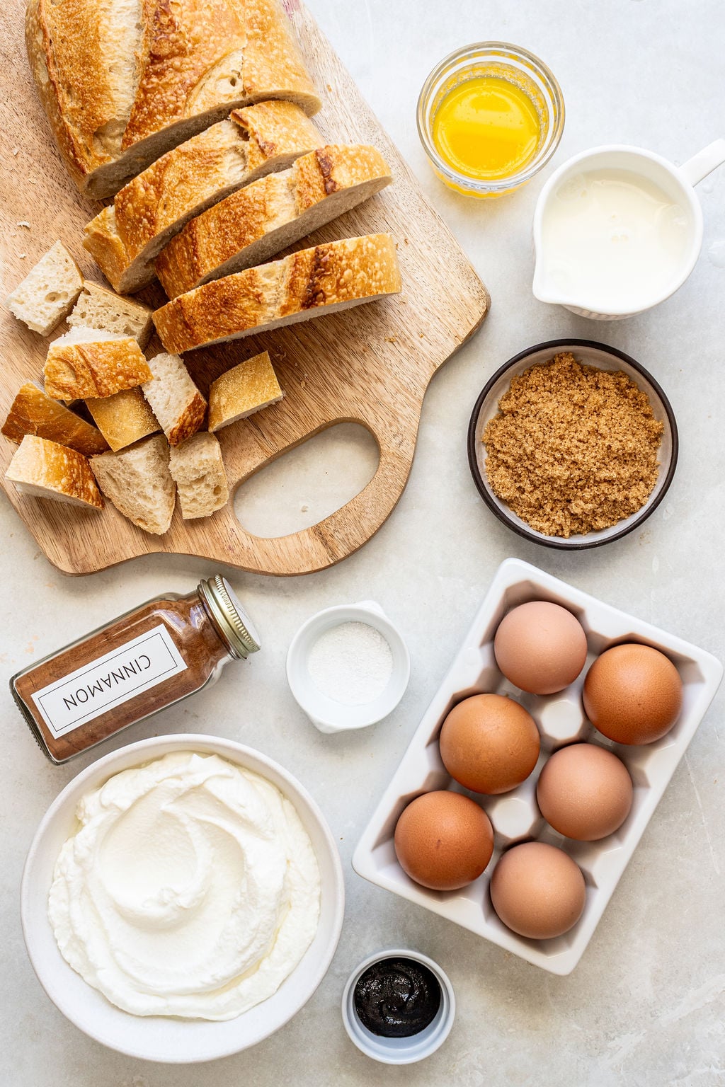 Ingredients for a recipe displayed: sliced bread, melted butter, cream, brown sugar, eggs, ricotta cheese, ground cinnamon, vanilla extract, and granulated sugar on a light surface.