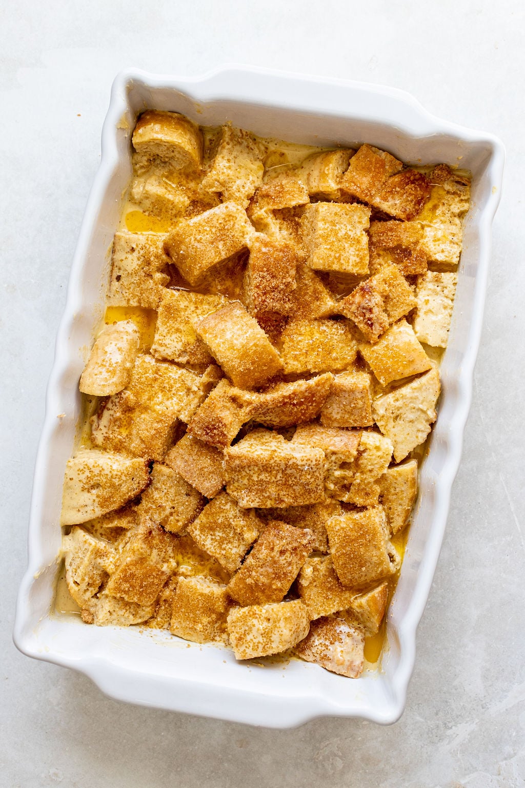 A white baking dish filled with cubed bread pieces, covered in a yellow liquid and sprinkled with brown sugar, prepared for baking.