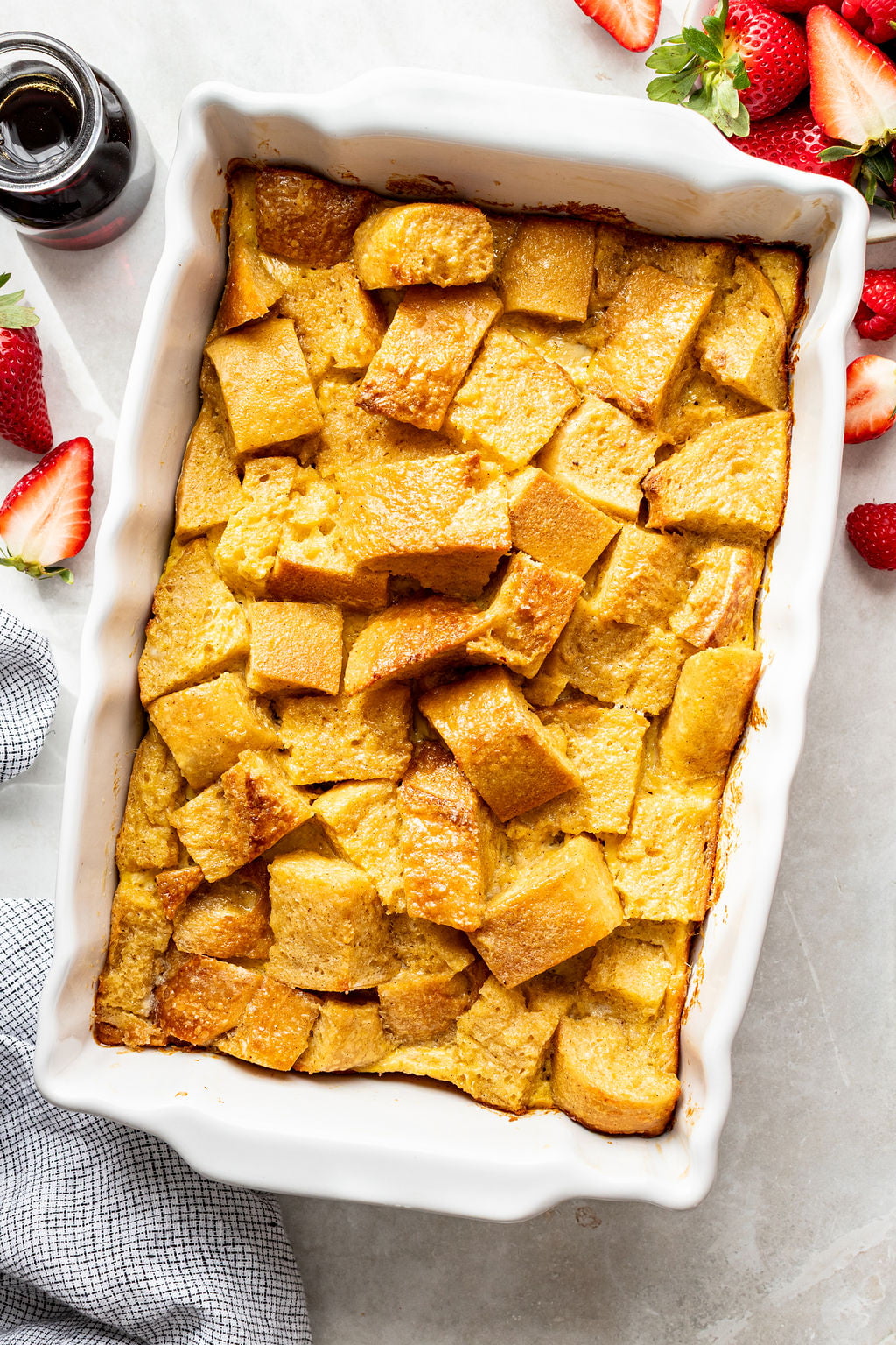 A rectangular white baking dish filled with golden-brown baked French toast casserole, with cut strawberries and a small syrup bottle nearby.
