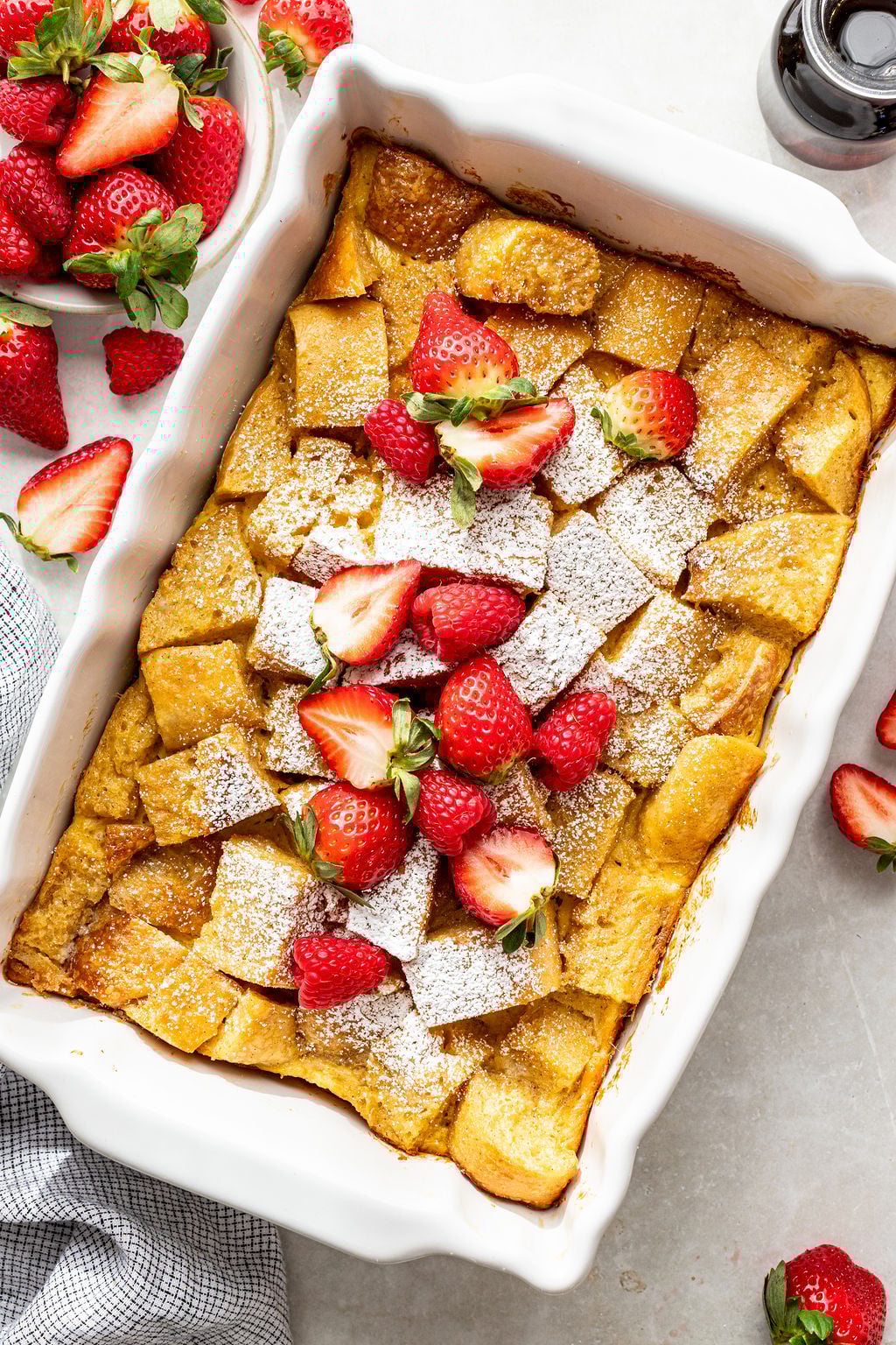 A baked French toast casserole topped with powdered sugar and fresh strawberries in a white baking dish, surrounded by whole strawberries.