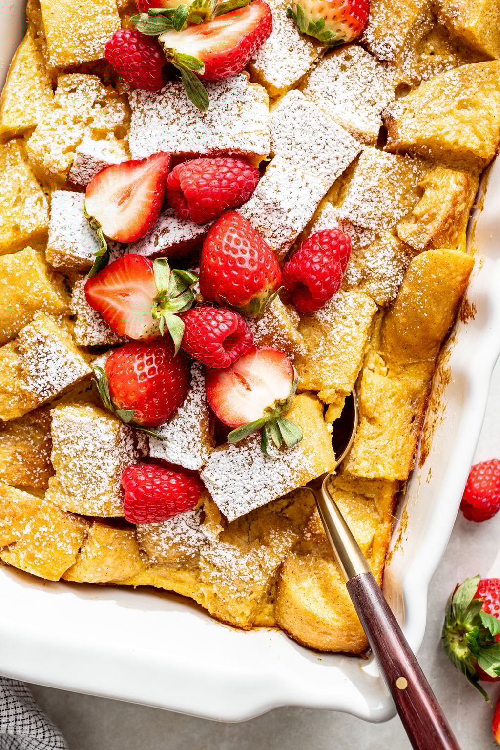 A baked bread pudding topped with powdered sugar and fresh strawberries, shown in a white dish with a serving spoon.