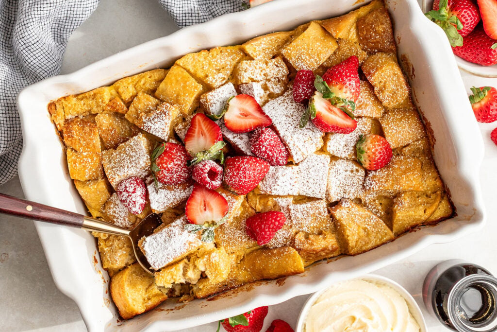 A baked French toast casserole topped with powdered sugar and fresh strawberries, with a serving spoon in the dish and a bowl of whipped cream nearby.