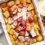 A baked French toast casserole topped with powdered sugar and fresh strawberries, with a serving spoon in the dish and bowls of syrup and whipped cream nearby.