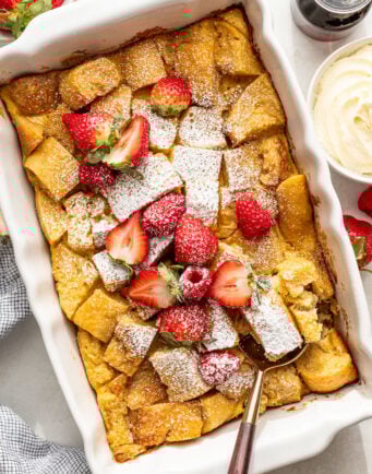 A baked French toast casserole topped with powdered sugar and fresh strawberries, with a serving spoon in the dish and bowls of syrup and whipped cream nearby.