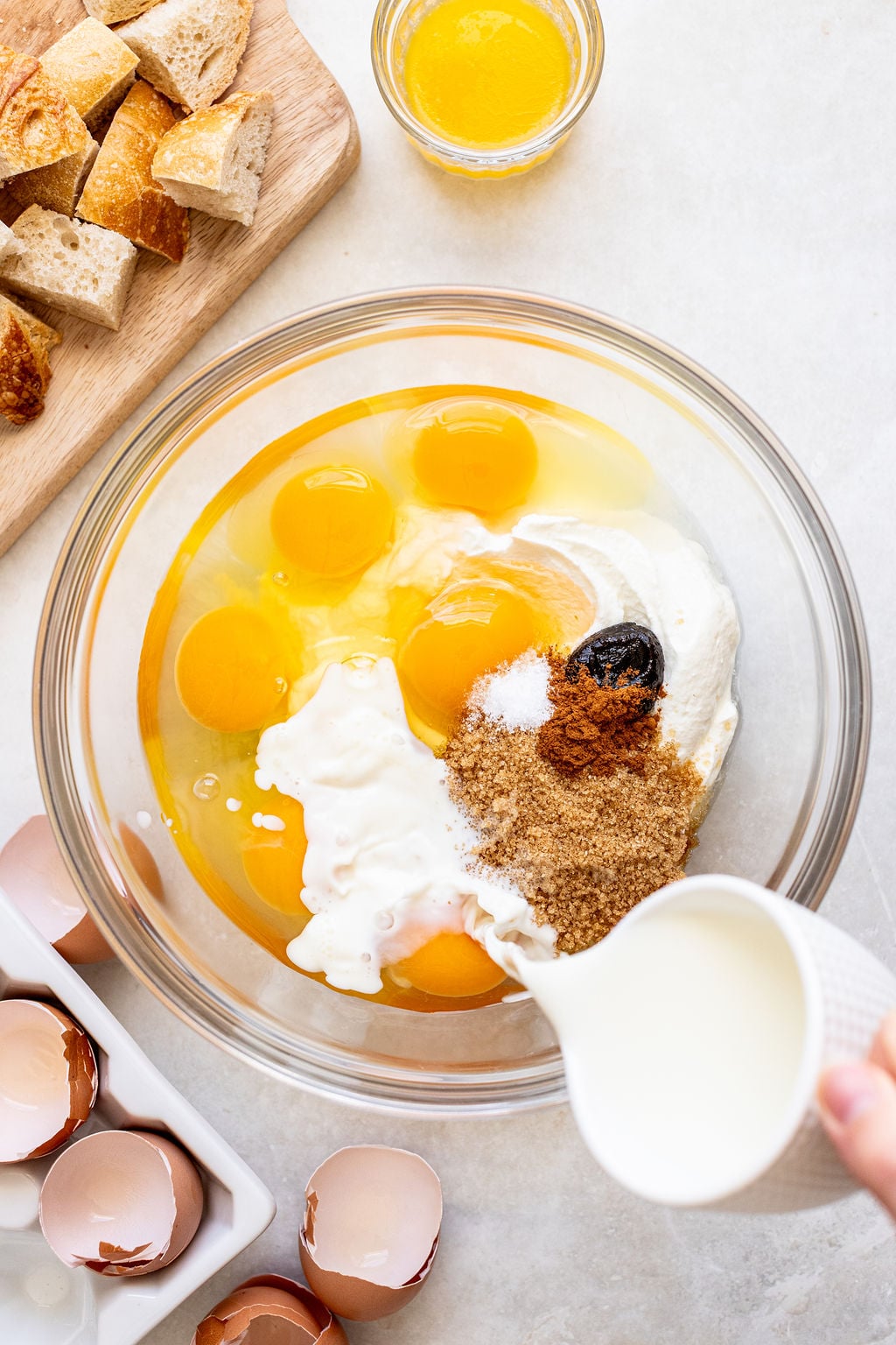A bowl with eggs, cream, sugar, cinnamon, and vanilla is being mixed with milk; broken eggshells, melted butter, and bread cubes are nearby on a counter.