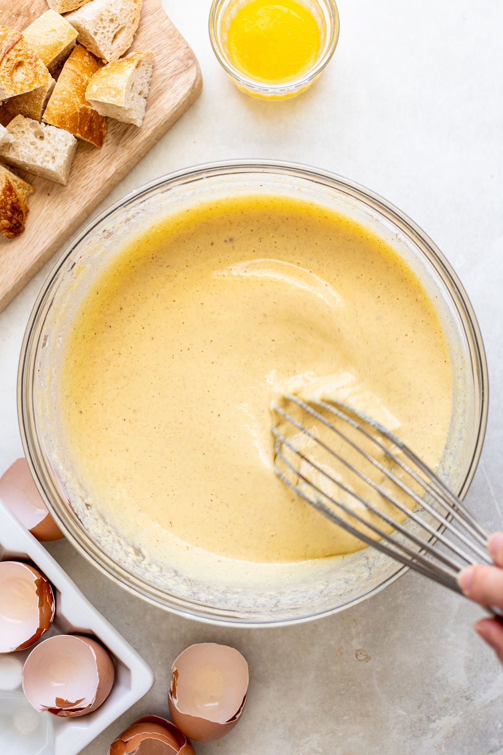 A hand whisks a bowl of yellow batter; egg shells, cubed bread, and melted butter are nearby on a light countertop.