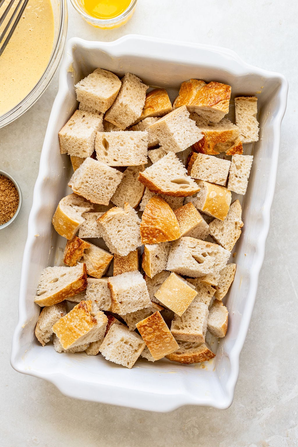Cubed bread pieces are arranged in a white baking dish, with a bowl of whisked liquid mixture and a small dish of brown sugar nearby.
