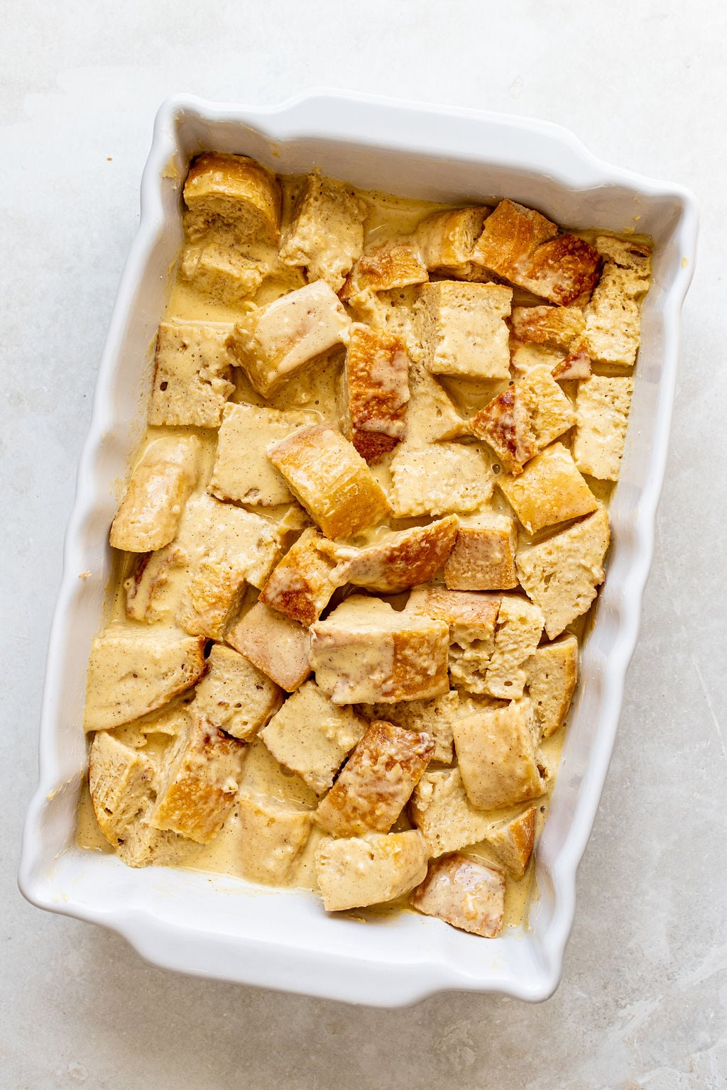 A white baking dish filled with cubed bread soaked in a creamy liquid, prepared for baking bread pudding or French toast casserole.