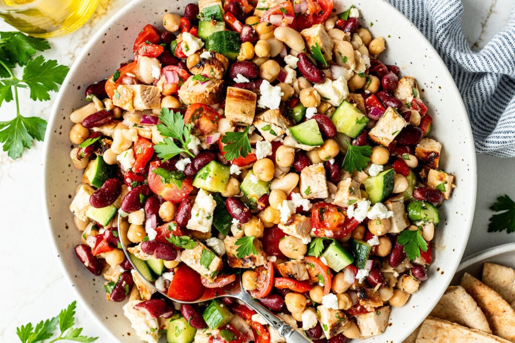 A bowl of Mediterranean salad with grilled chicken, chickpeas, kidney beans, cucumber, cherry tomatoes, parsley, and feta cheese.